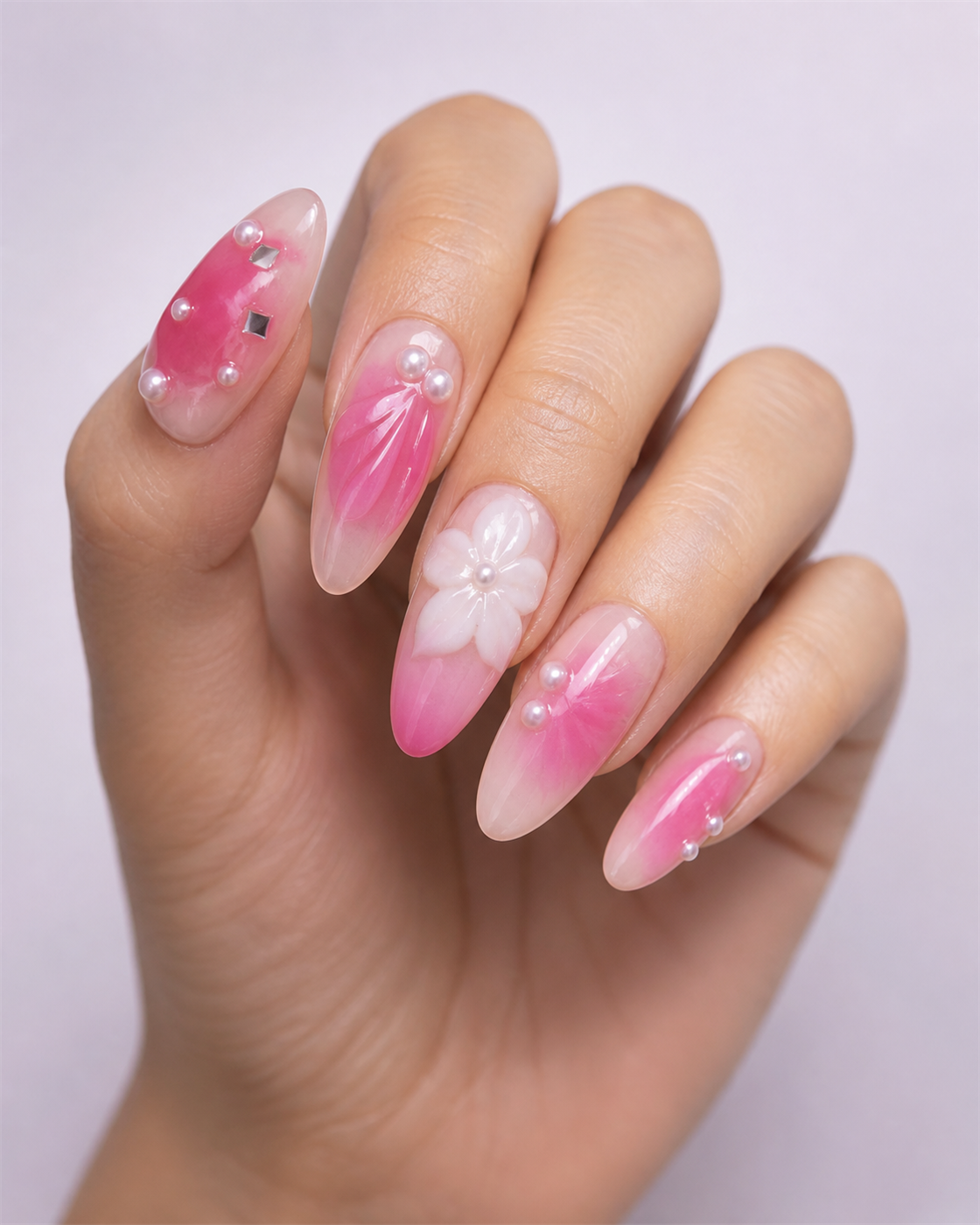 Hand wearing pink gradient almond press-on nails with white flowers, pearls and reflective accents.